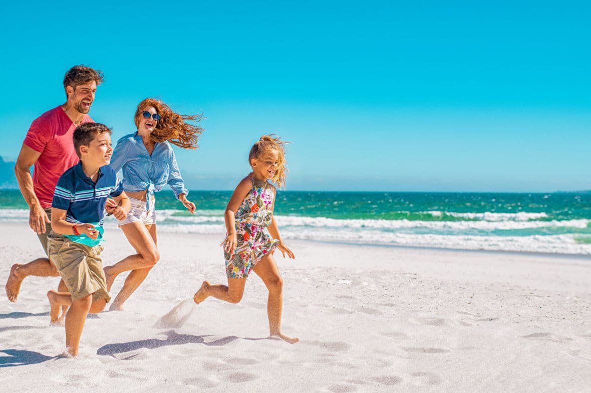 Família feliz em férias na praia, vestindo roupas brancas e chapéus de sol, com criança segurando uma boia amarela